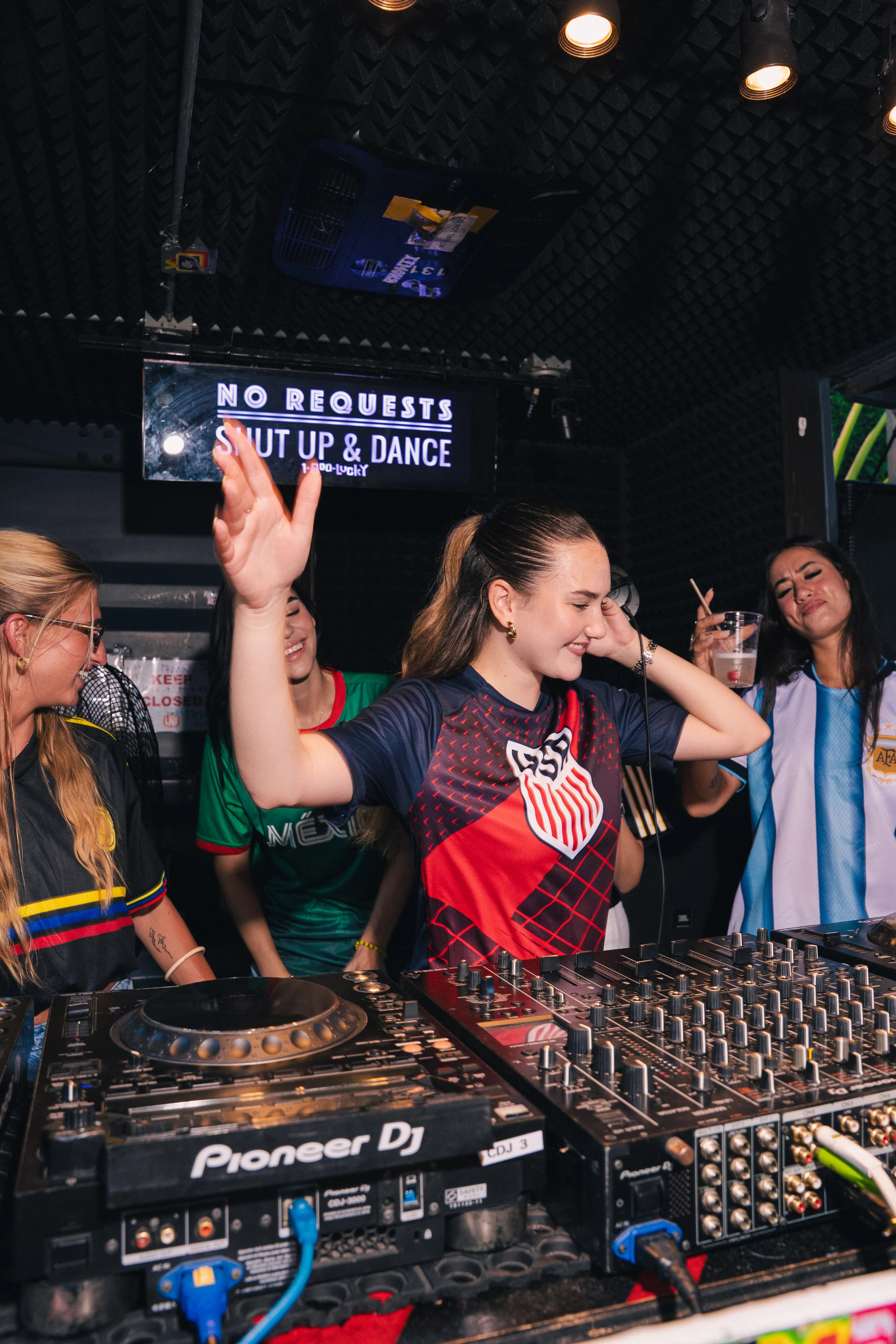 A World Cup-season night at 1-800-Lucky — a DJ in a red-and-navy jersey throws a hand up at the decks, surrounded by fans in Colombia, Mexico, and Argentina jerseys beneath the 'No Requests, Shut Up & Dance' sign.
