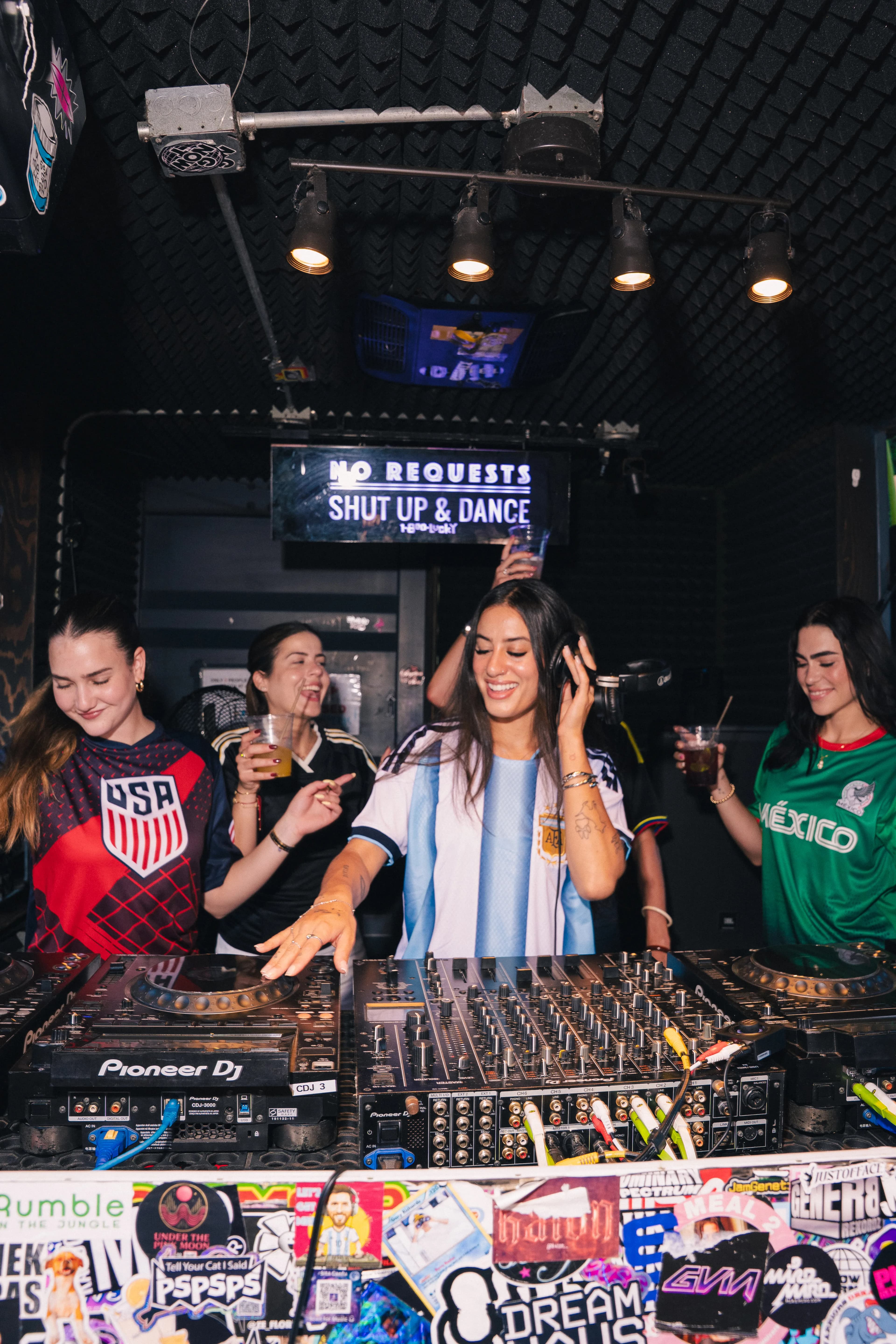 A World Cup-season night at 1-800-Lucky — a DJ in a light-blue Argentina jersey mixes on Pioneer decks between fans in USA and Mexico jerseys, over a sticker-covered booth.