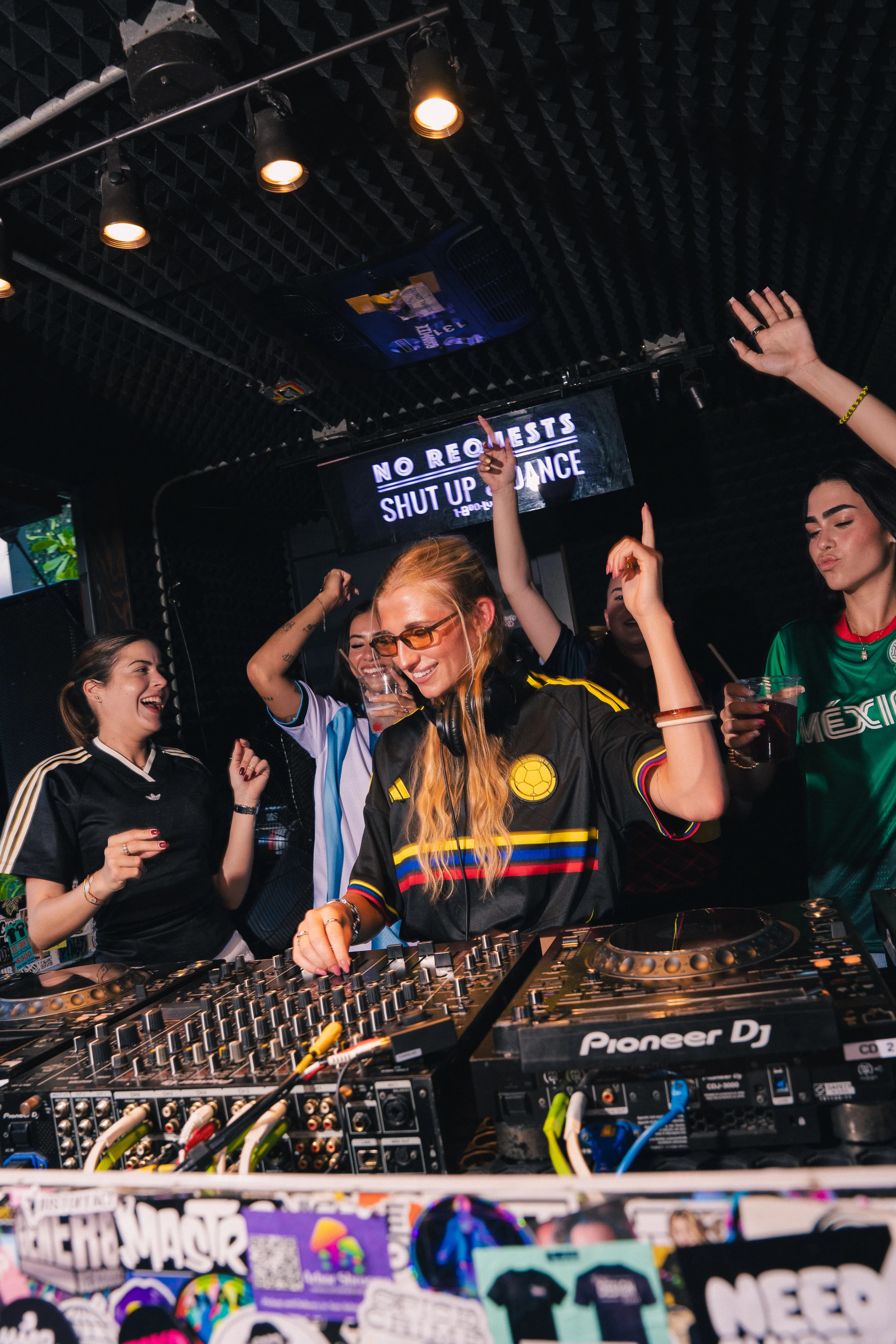 A World Cup-season night at 1-800-Lucky — a DJ in a black Colombia jersey works the decks as a jersey-clad crowd dances with hands raised under the 'No Requests, Shut Up & Dance' sign.