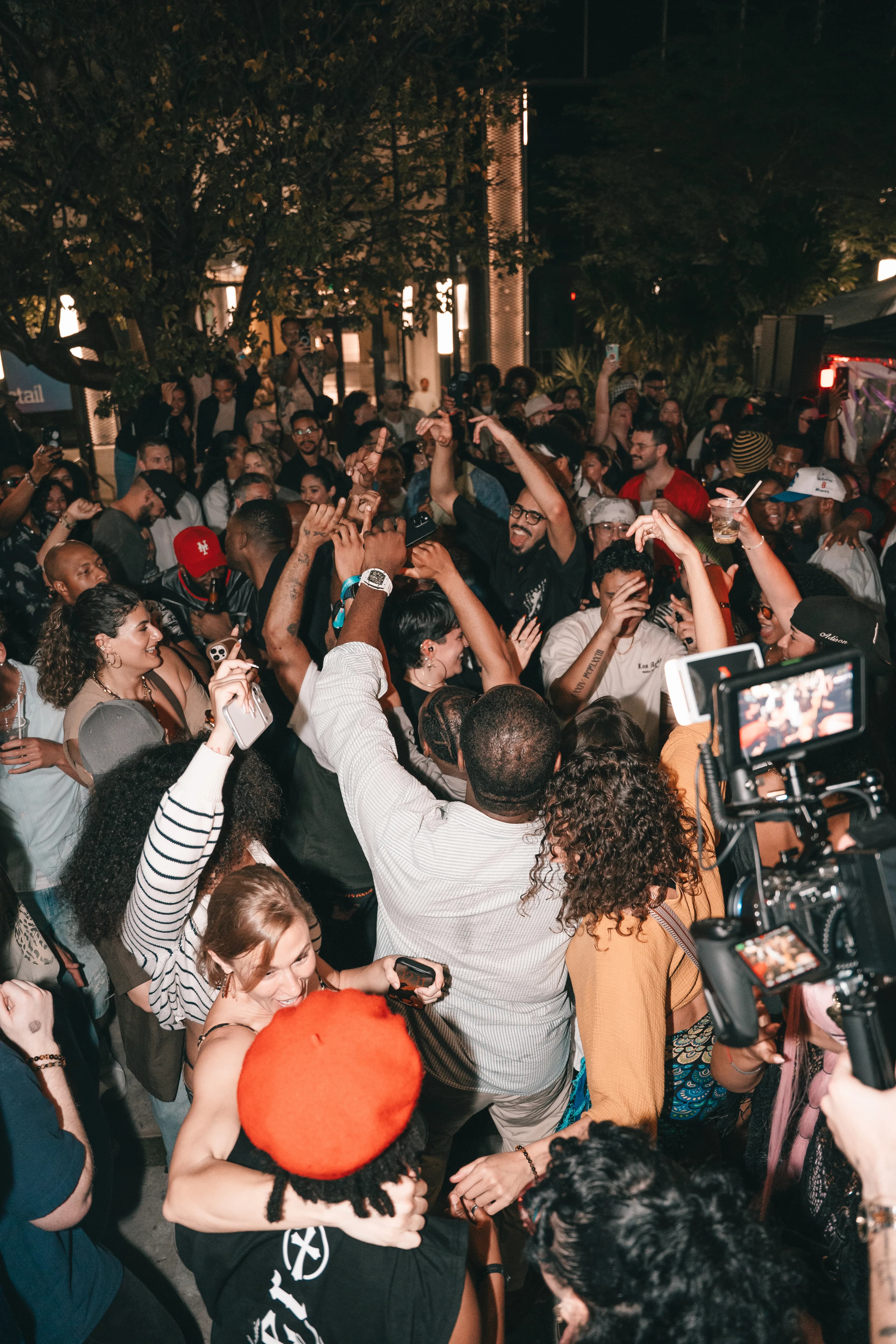 A packed nighttime crowd dances with hands raised at an outdoor Dante's HiFi+ event, a camera monitor glowing at the edge of the frame.