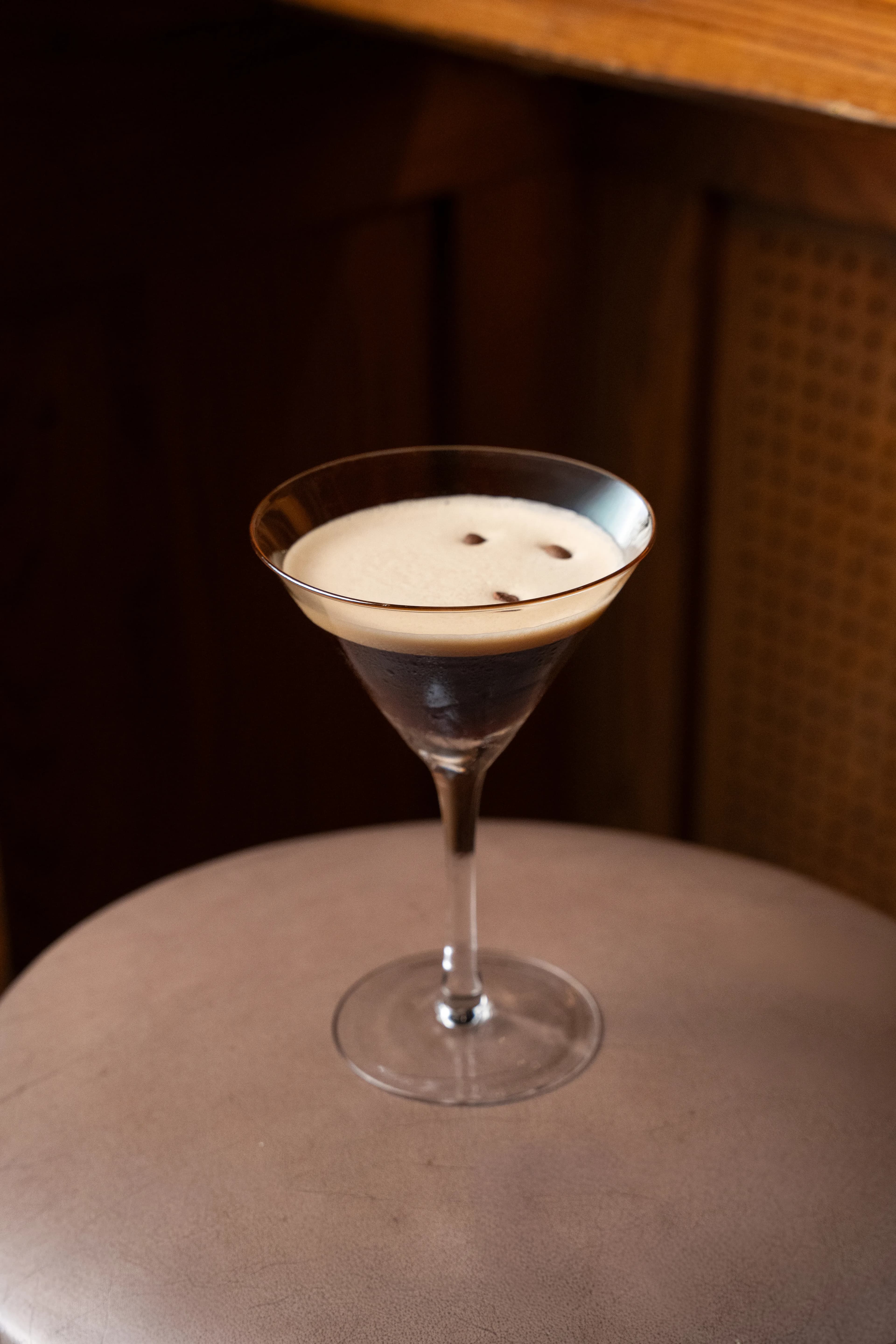 An espresso martini topped with three coffee beans rests on a leather stool against warm wood paneling at Dante's HiFi+.