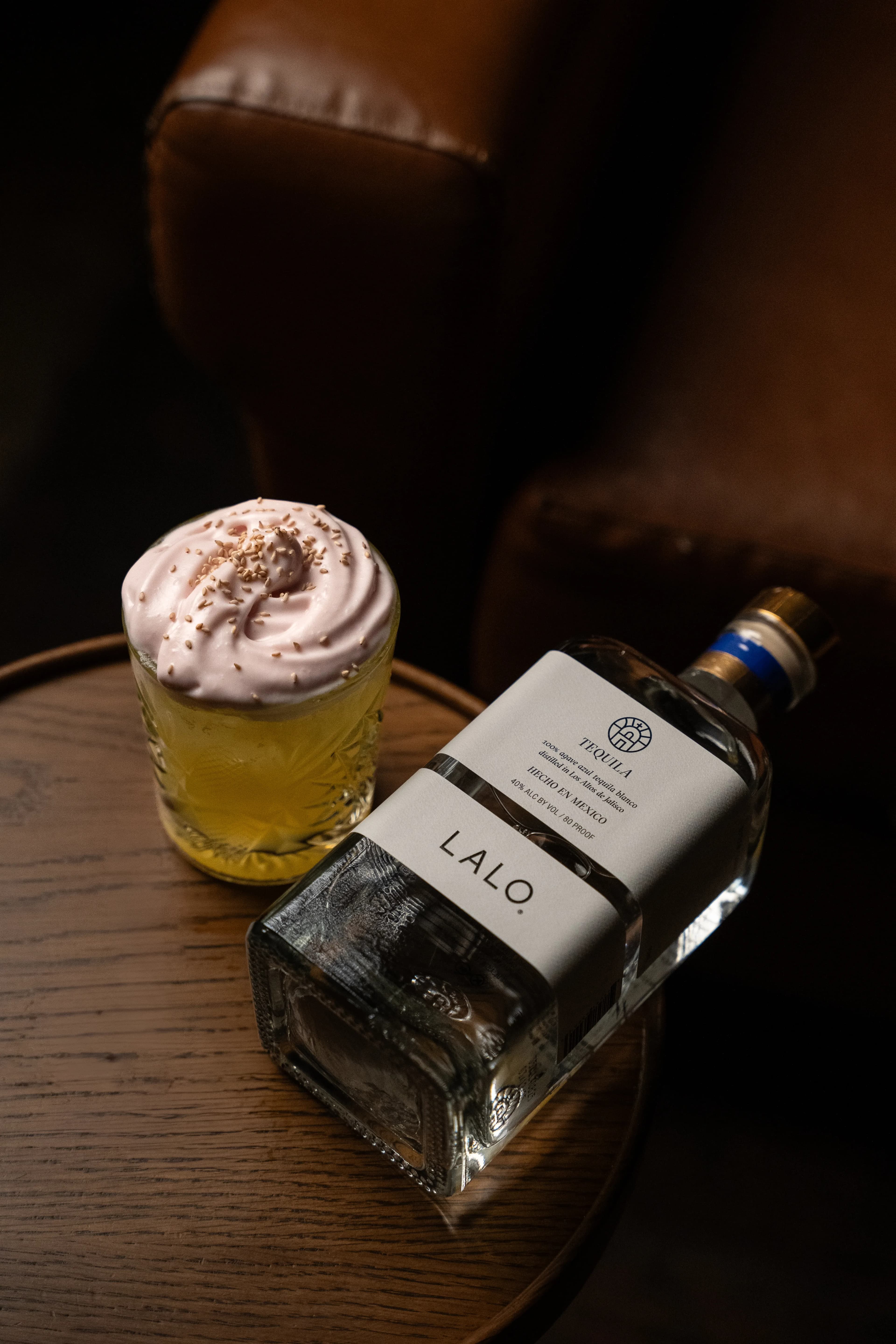 A pink-foam cocktail dusted with sesame beside a bottle of Lalo tequila on a round wooden table at Dante's HiFi+.