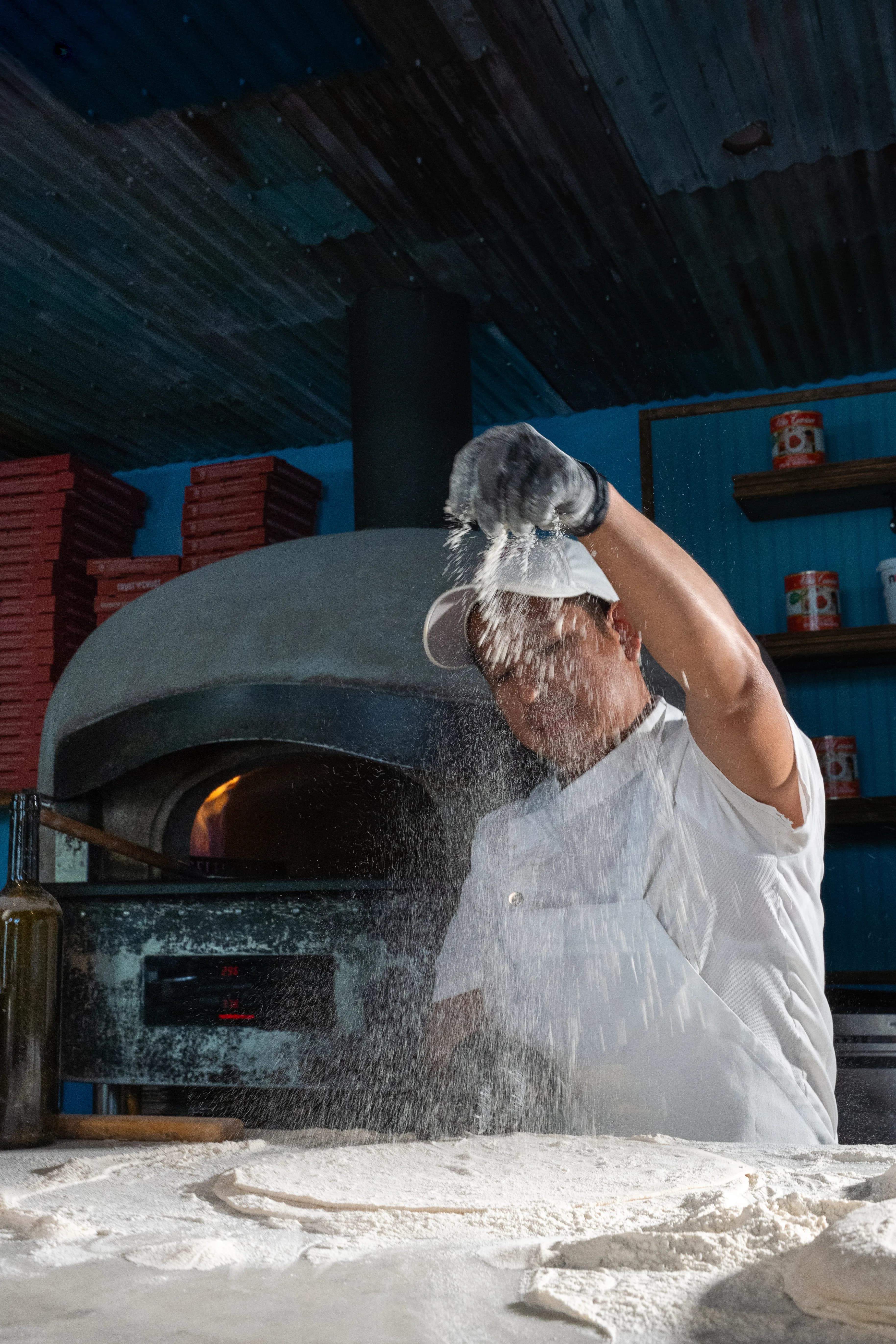 A pizzaiolo tosses floured dough before a blue-tiled wood-fired oven, flour catching the light at DC Pie Co.