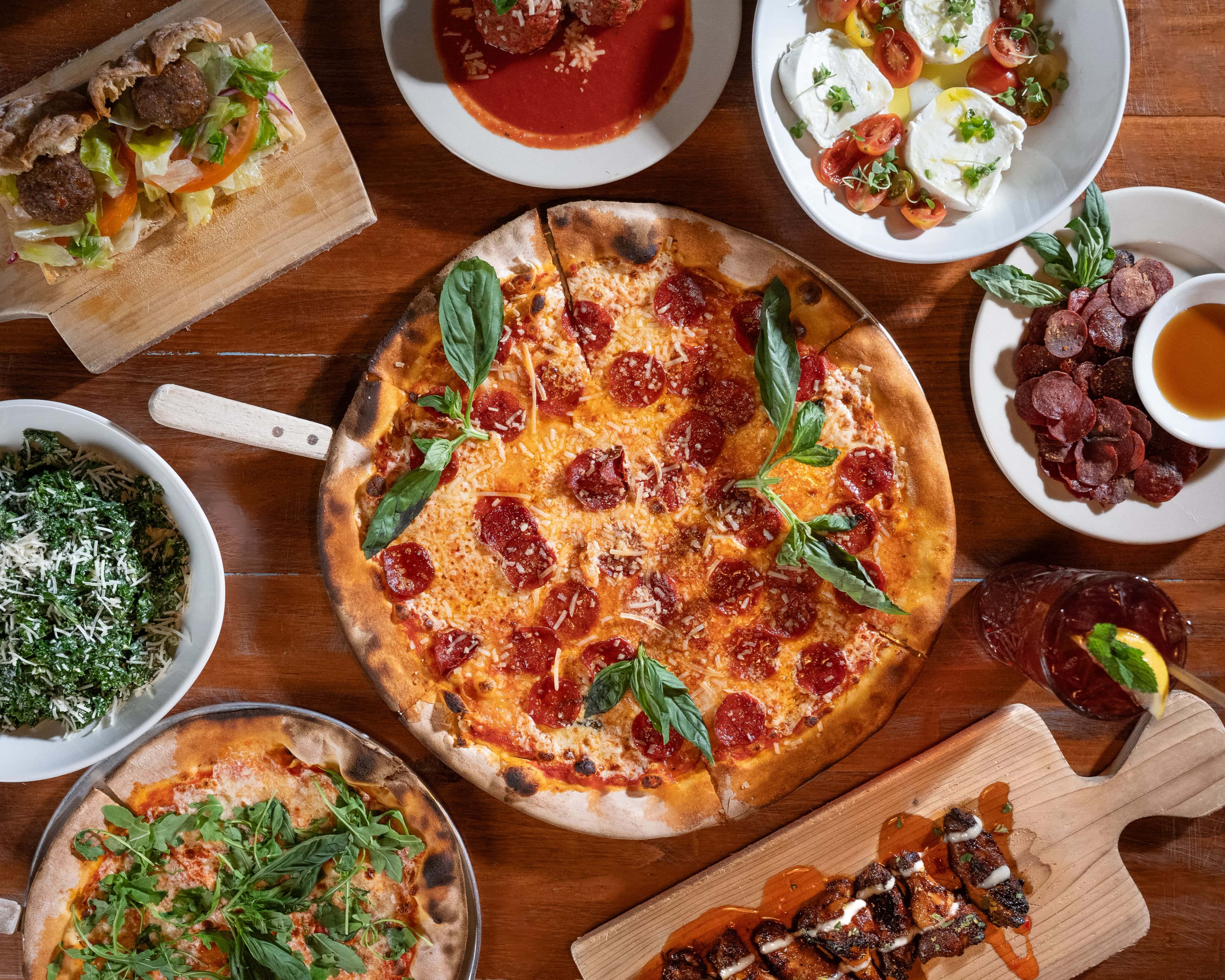 An overhead spread of DC Pie Co. dishes around a large pepperoni-and-basil brick-oven pizza — meatballs, burrata, charcuterie, pasta, salad, and skewers on a wood table.