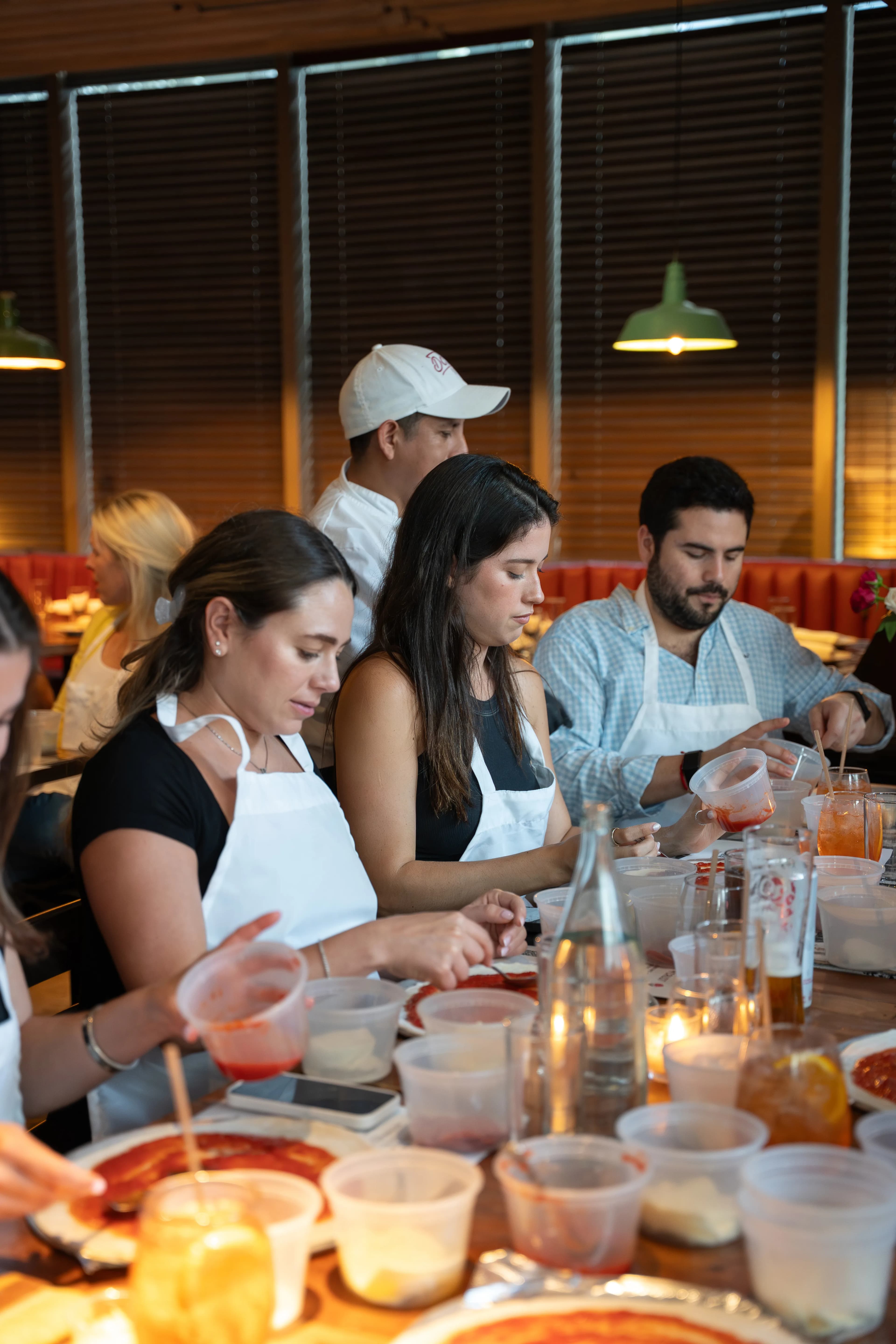 Guests in aprons assemble pizzas at a long candlelit table during a DC Pie Co. pizza-making class.