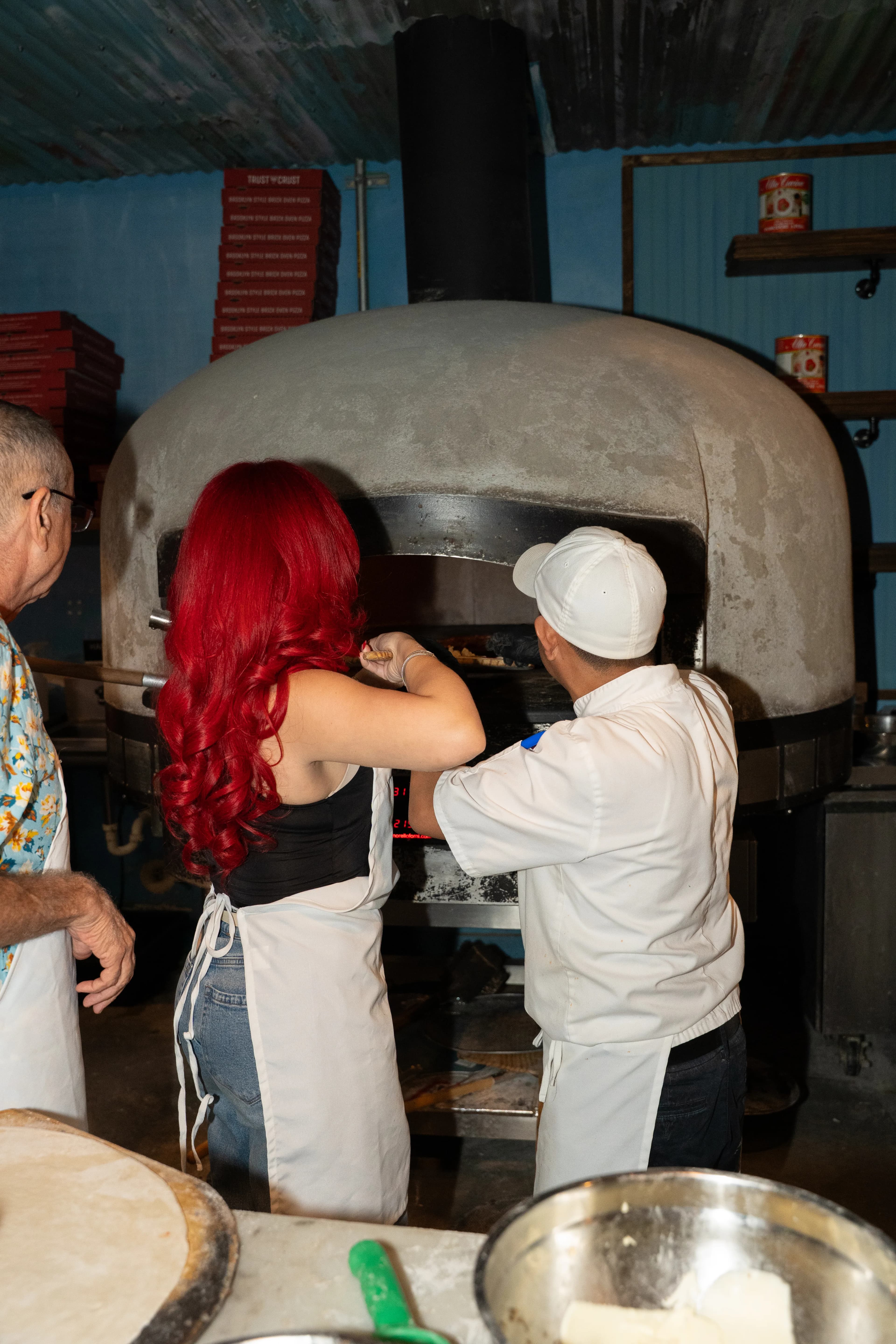 A red-haired guest and a pizzaiolo slide a pizza into a rustic wood-fired oven at DC Pie Co.