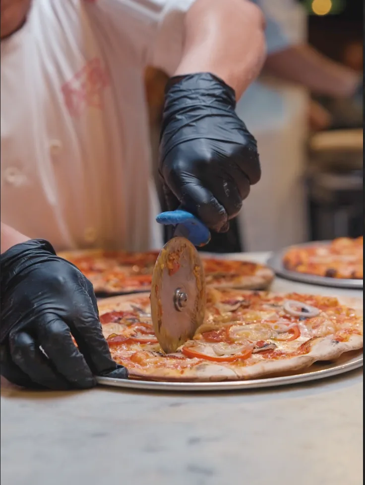 Gloved hands cut a fresh pizza with a wheel on the marble counter at DC Pie Co.