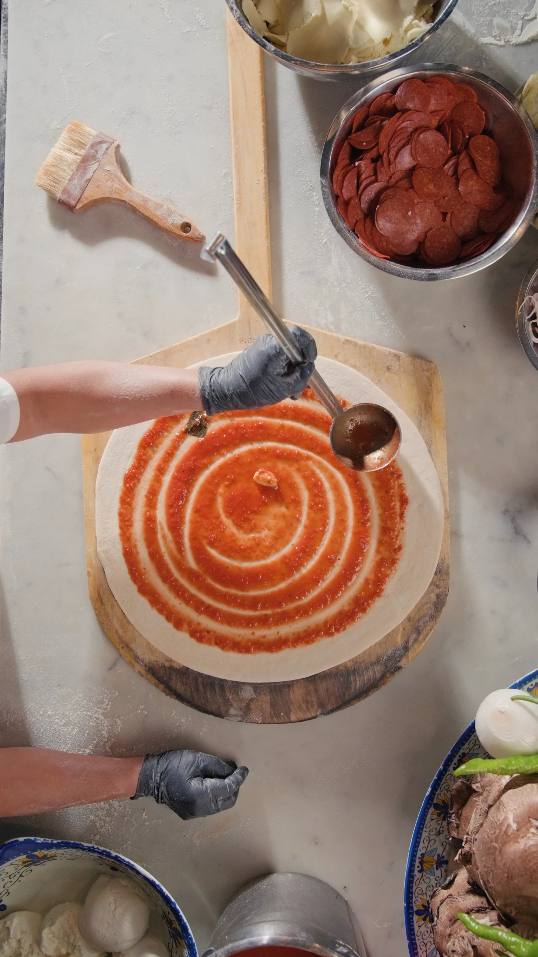 Overhead of gloved hands spiraling sauce onto fresh dough surrounded by pepperoni and topping bowls at DC Pie Co.