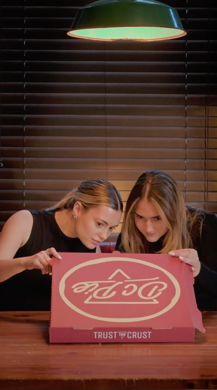Two guests lean in to open a pink DC Pie Co. pizza box reading 'Trust the Crust' under a warm pendant lamp.