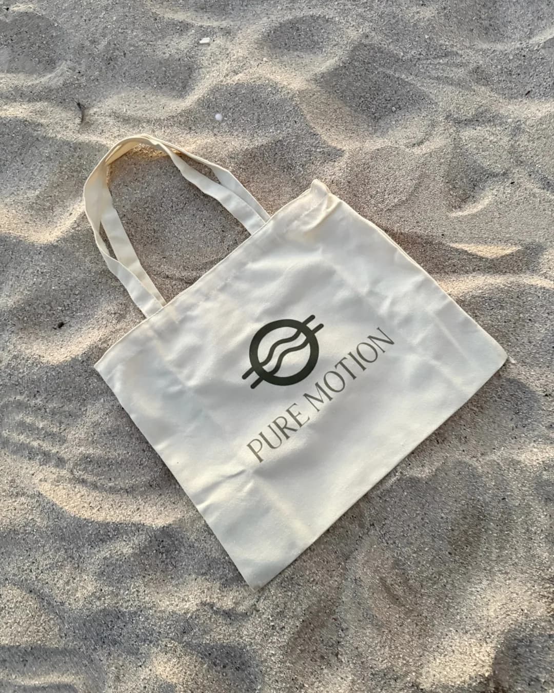 A white Pure Motion logo tote bag resting on the sand.