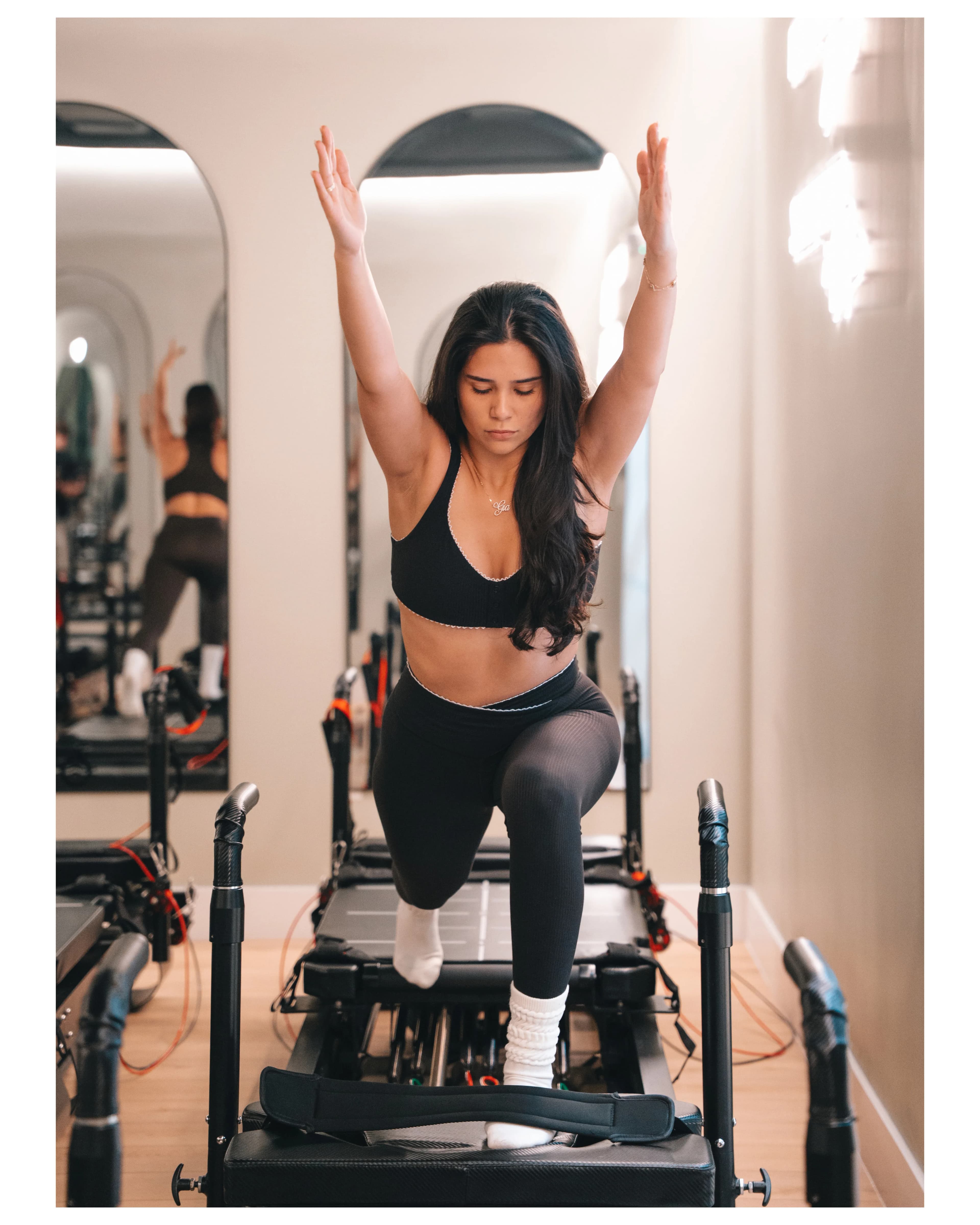 A woman reaches overhead on a reformer in a sunlit Pure Motion studio.