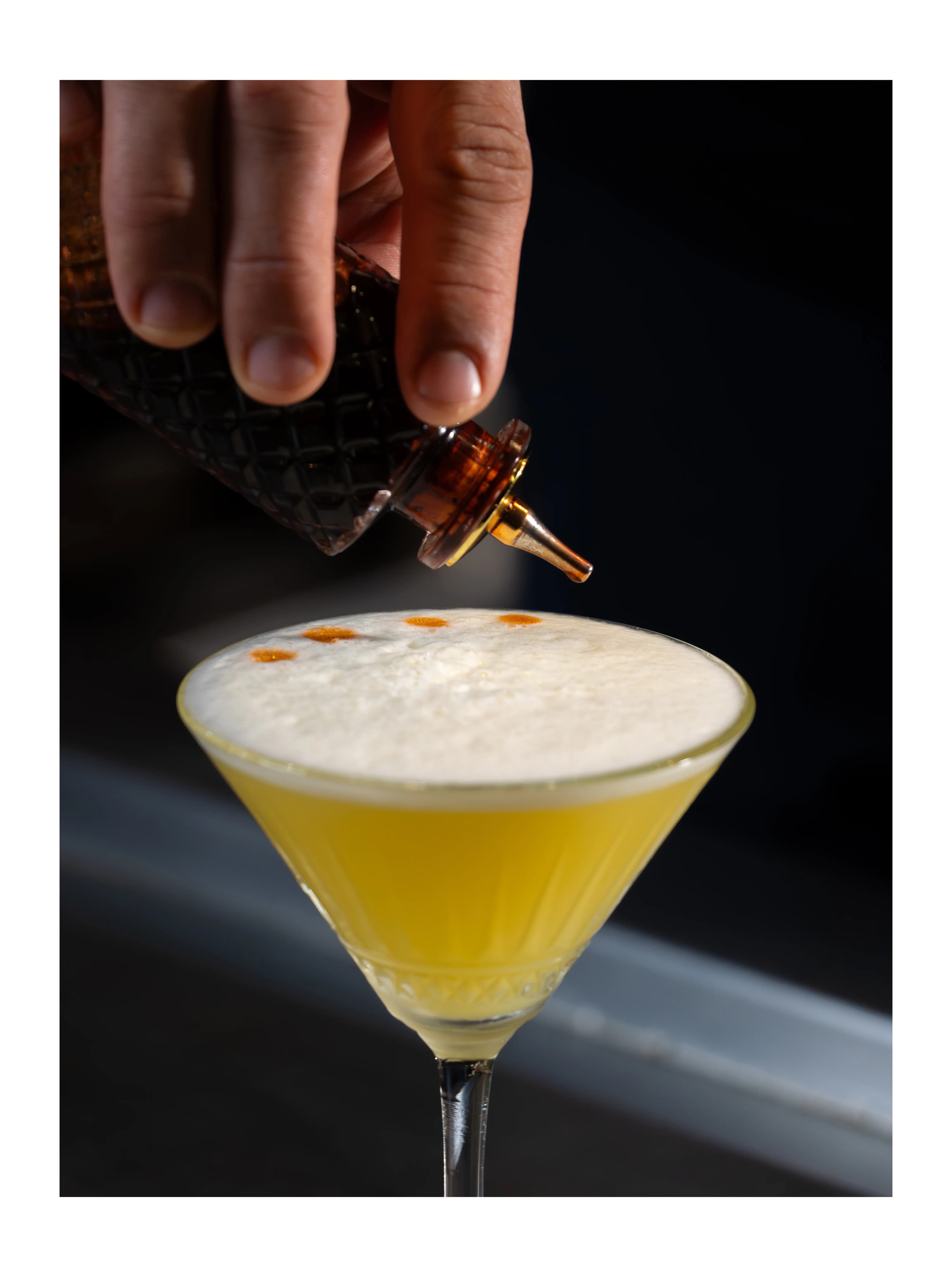A hand drops bitters over a frothy citrus cocktail in a coupe glass at Sushi Sake.