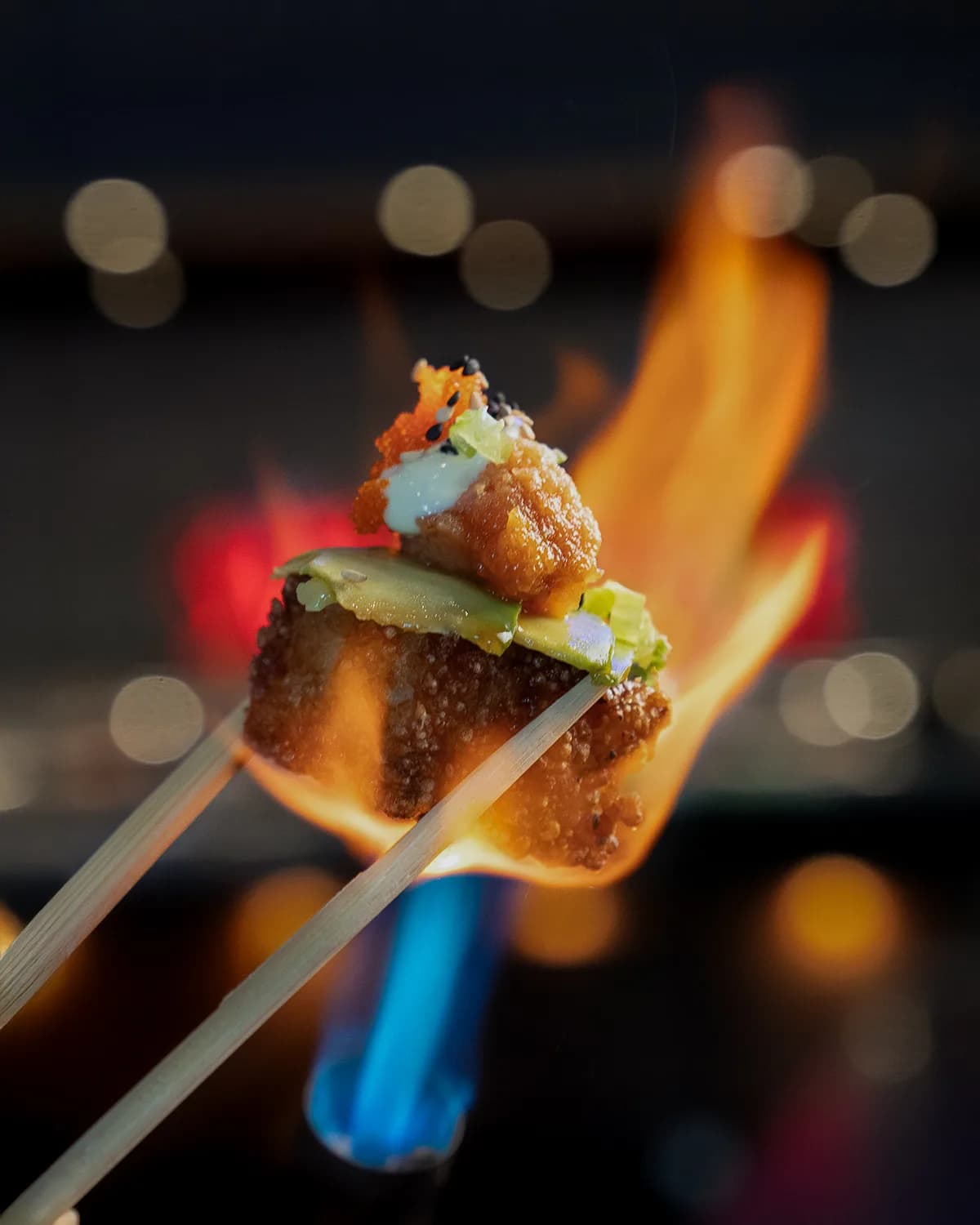 Chopsticks hold a torched crispy-rice bite topped with fish over an open flame at Sushi Sake.