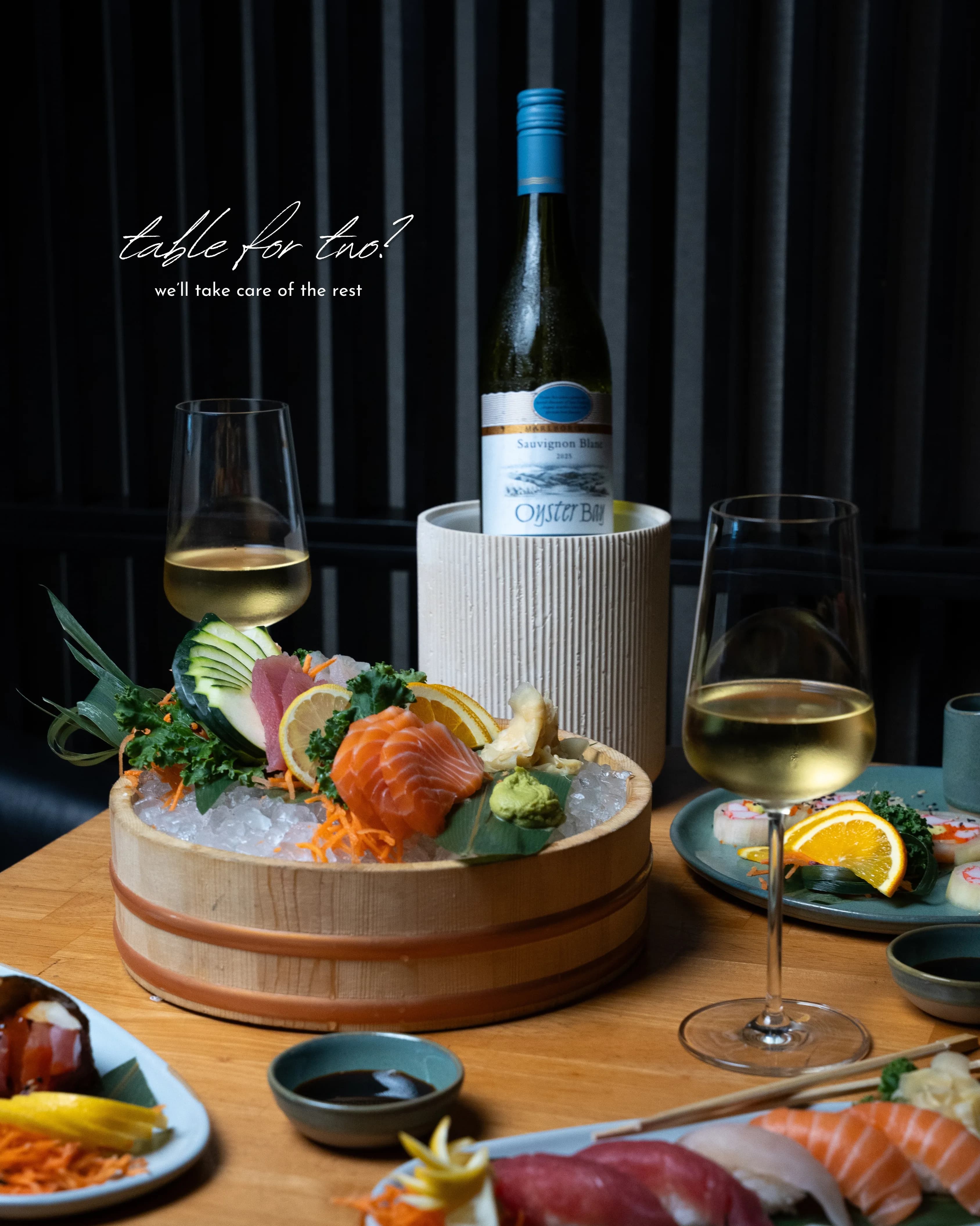 A Sushi Sake 'table for two' image — a sashimi boat on ice beside a sake bottle and wine glasses.