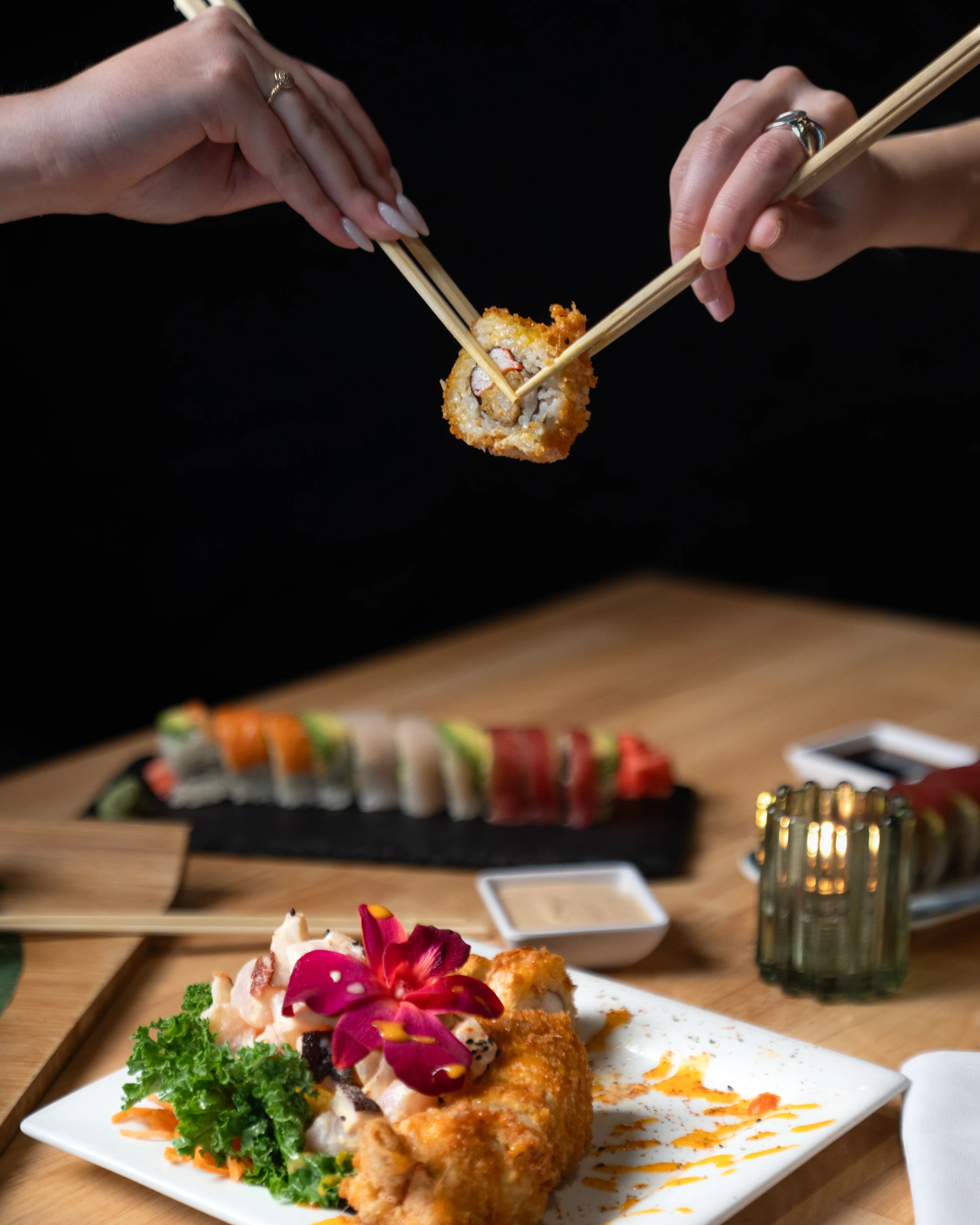 Chopsticks lift a crispy bite above a plated dish and a sushi roll at Sushi Sake.