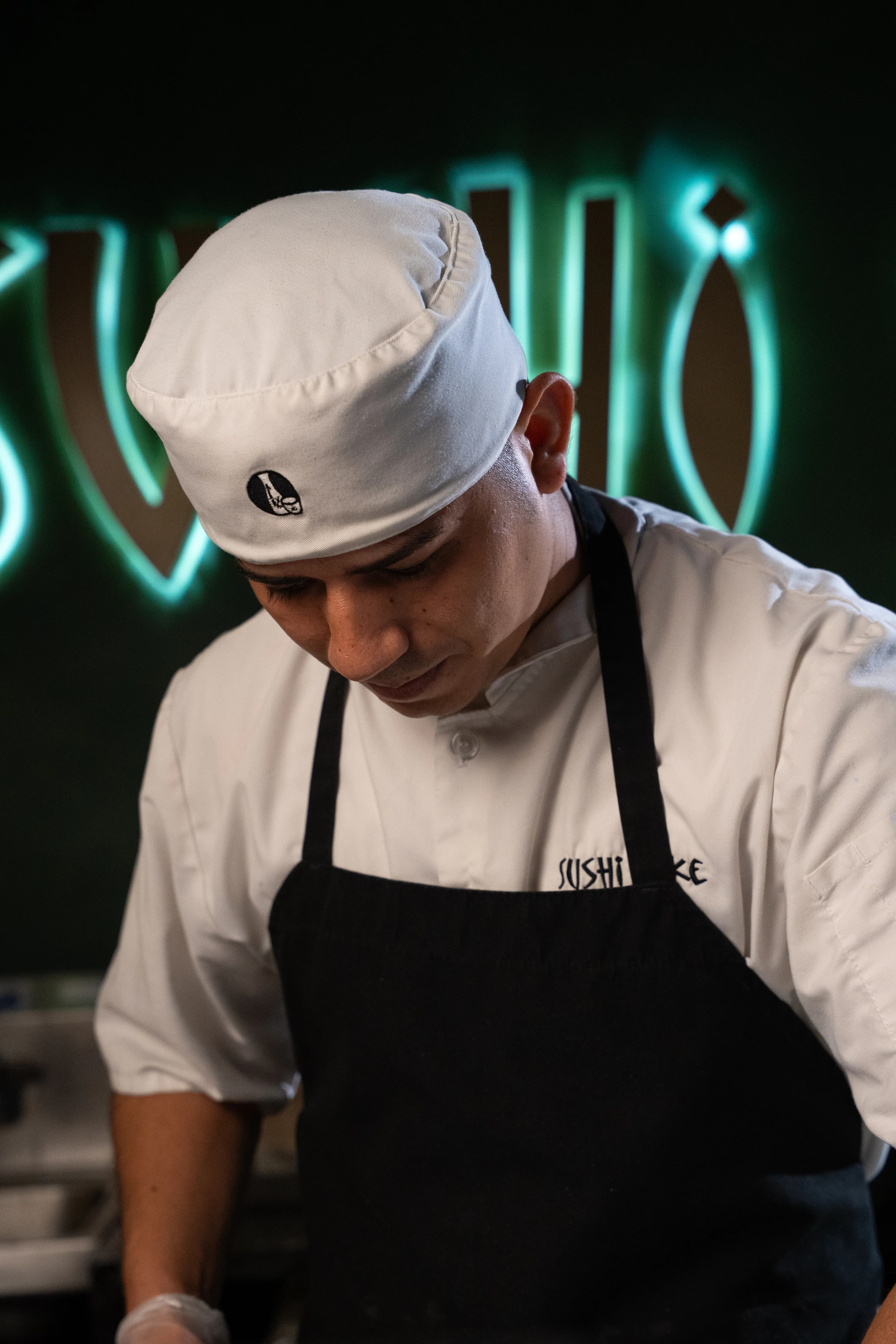 A Sushi Sake chef in cap and apron works at the bar beneath the neon sign.