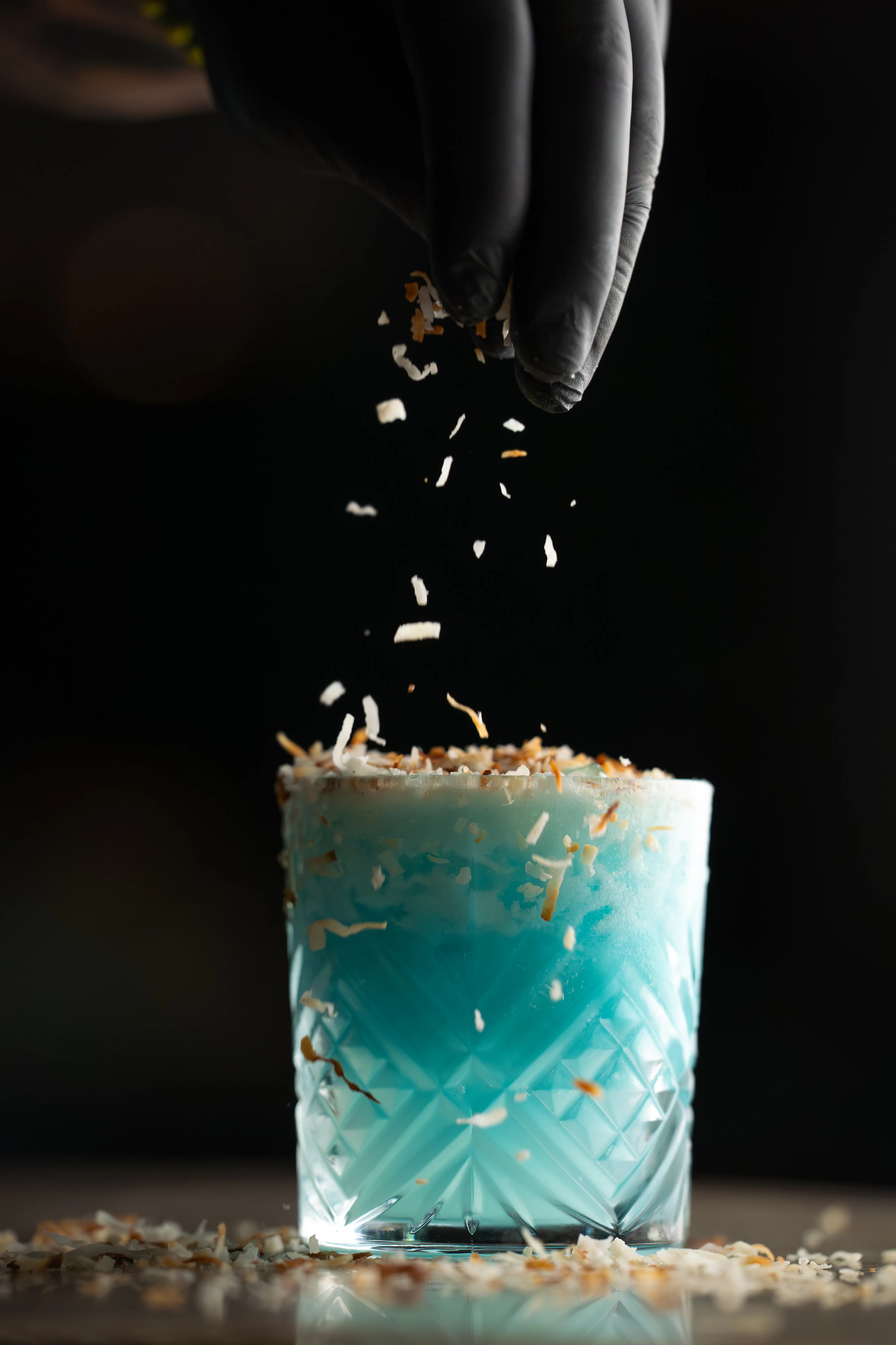 A gloved hand sprinkles garnish over a bright blue cocktail at Sushi Sake.