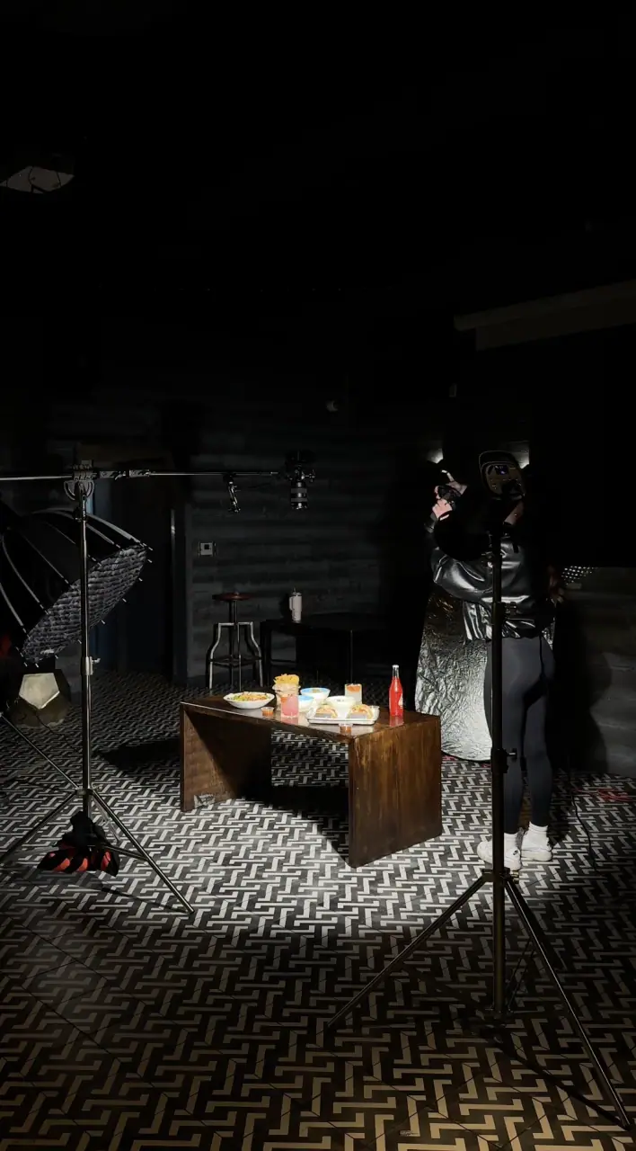 A dim studio setup at Coyo Taco — a small wood table of lit candles and hot sauce lit by a standing softbox as a crew member adjusts a light on a patterned floor.
