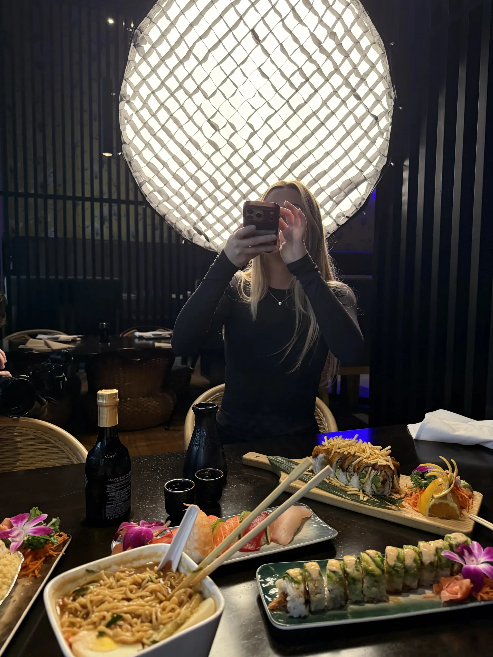 A guest photographs a spread of Sushi Sake rolls beneath a glowing round fixture.